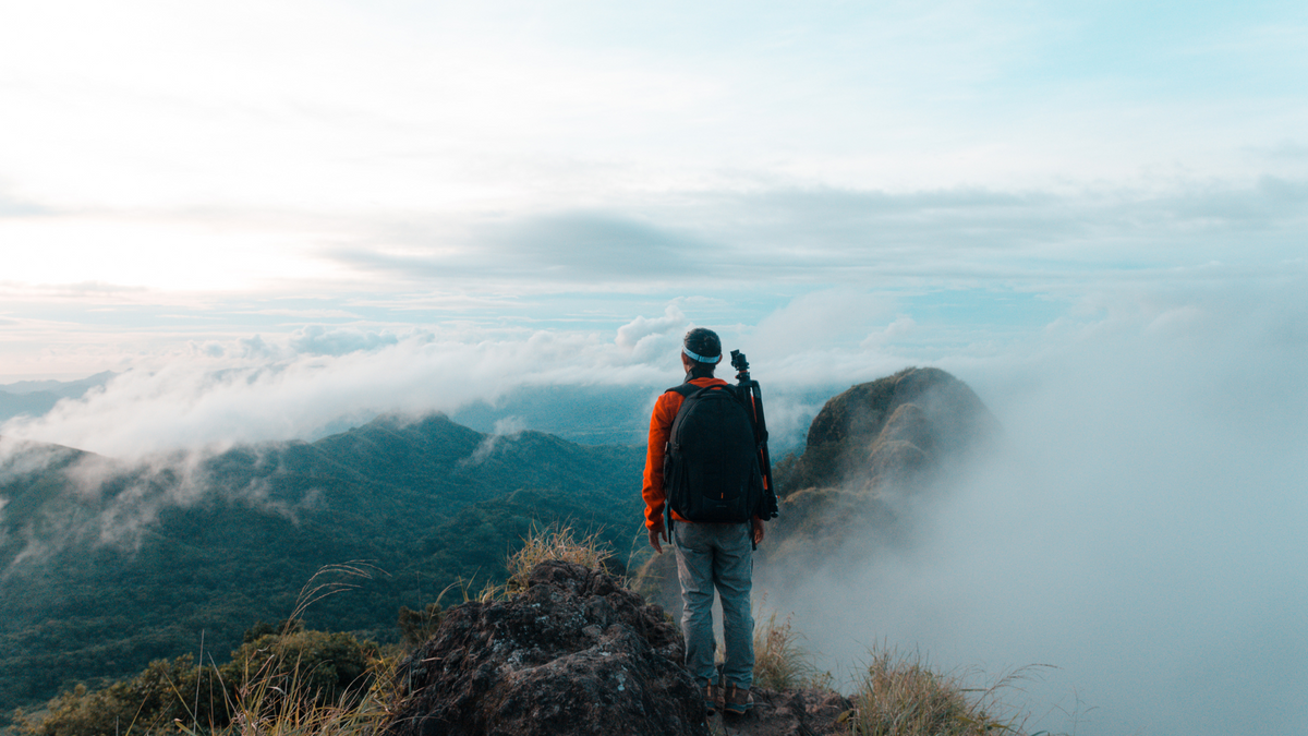 Person standing on a mountain peak with a backpack and camera, overlooking clouds.